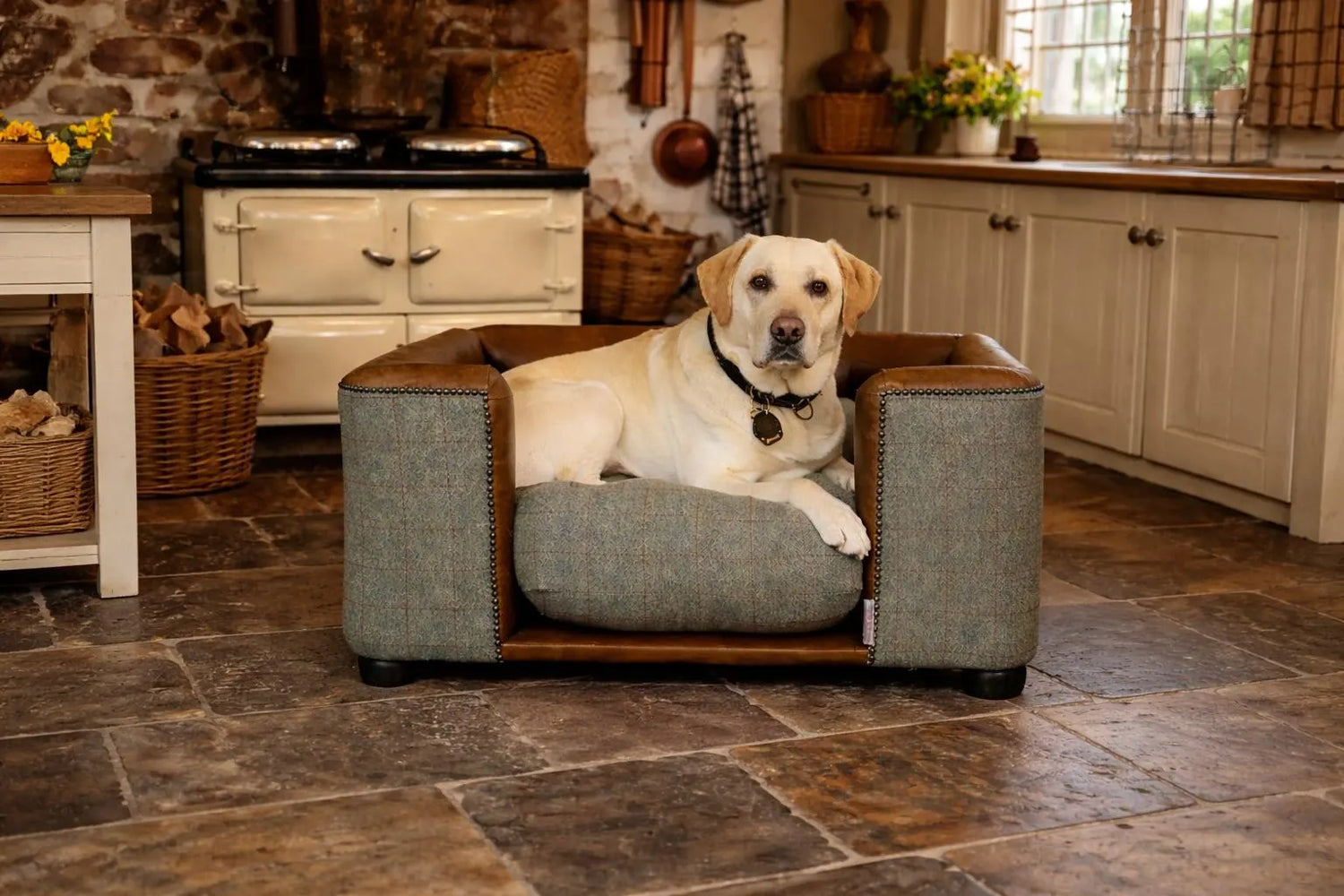 A golden Labrador lies in a luxury dog sofa in front of an Aga in a country kitchen. The bed has soft tan leather inside, brown Harris Tweed outside, bronze stud detailing, and sits on mahogany bun feet.
